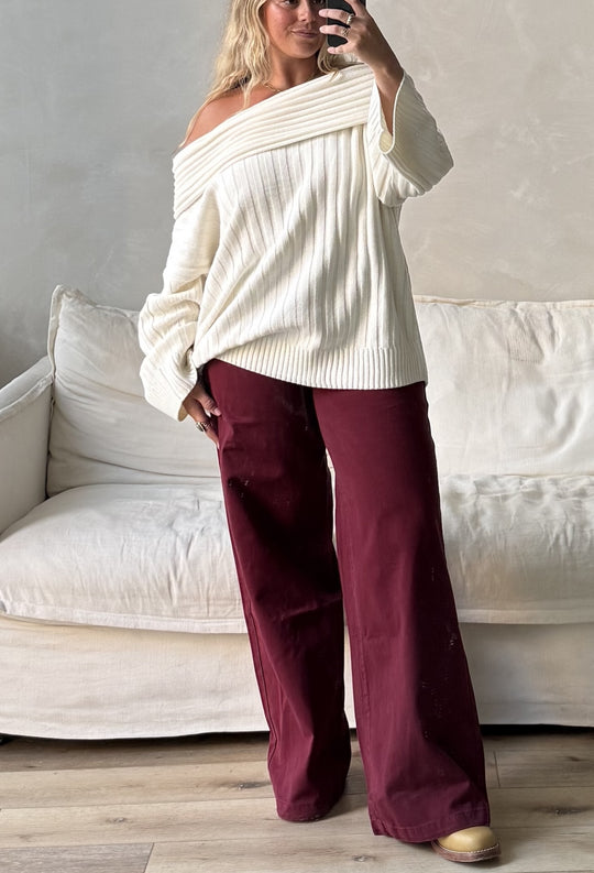 Woman taking a mirror selfie wearing WeWoreWhat white off the shoulder sweater with low rise baggy maroon pants with white couch in background