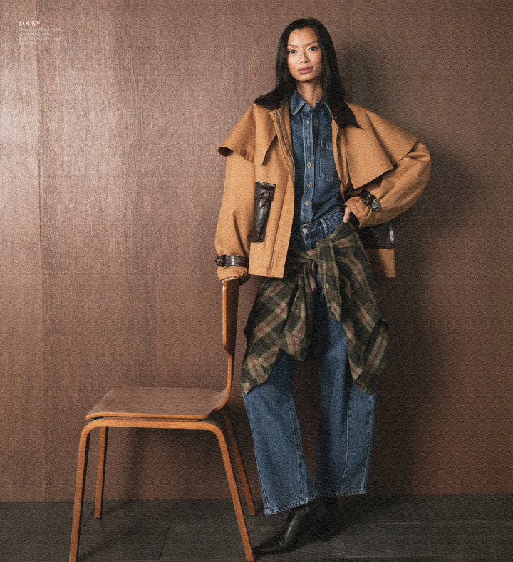 Woman in a stylish outfit with a tan coat and plaid shirt, standing against a brown background.
