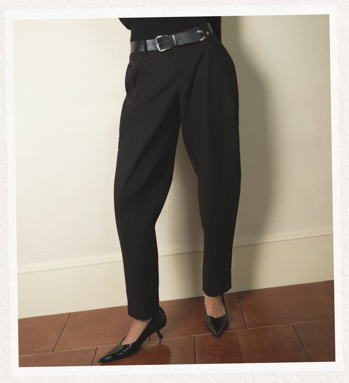 Person wearing black dress pants and shoes against a plain wall.