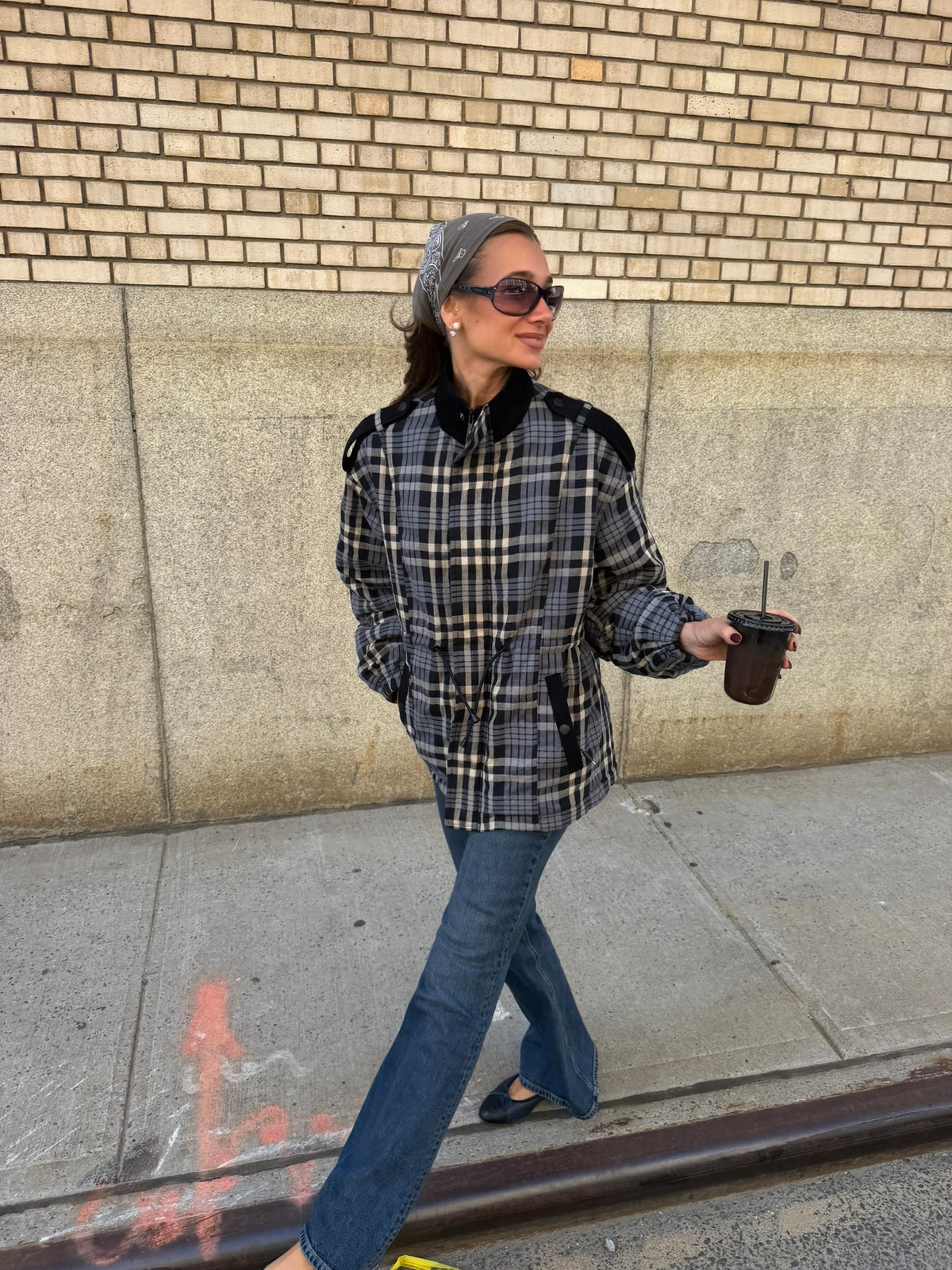 A woman walking on a city sidewalk wearing a black, gray, and beige plaid jacket with black shoulder panels, blue jeans, and holding a brown coffee cup.