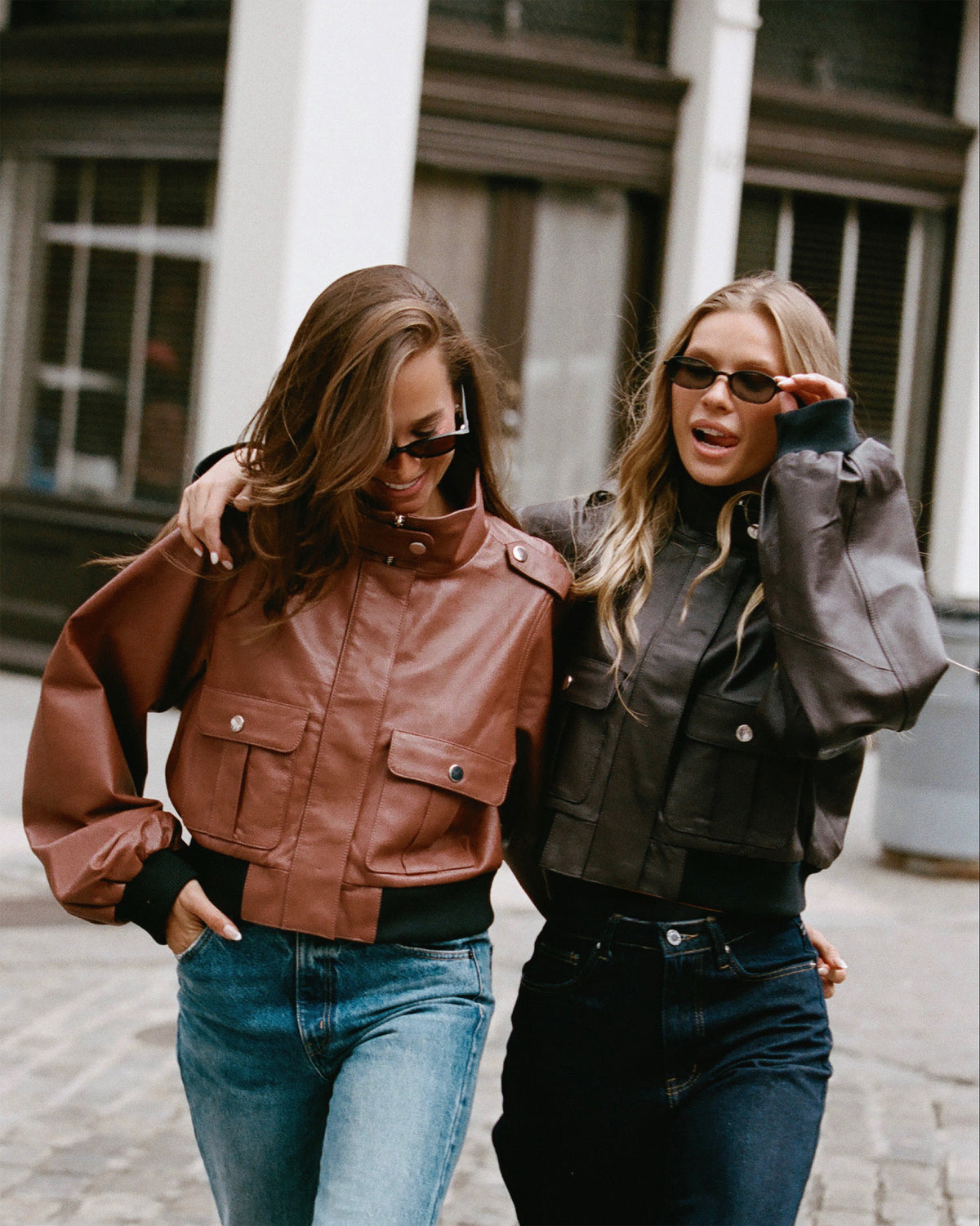 Two women walking arm-in-arm on a city street, each wearing a cropped leather jacket. The woman on the left wears a brown leather jacket, and the woman on the right wears a dark brown leather jacket.