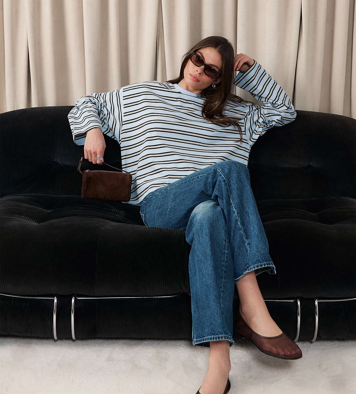 Woman sitting on a black sofa wearing a striped sweater and jeans, holding a brown handbag.