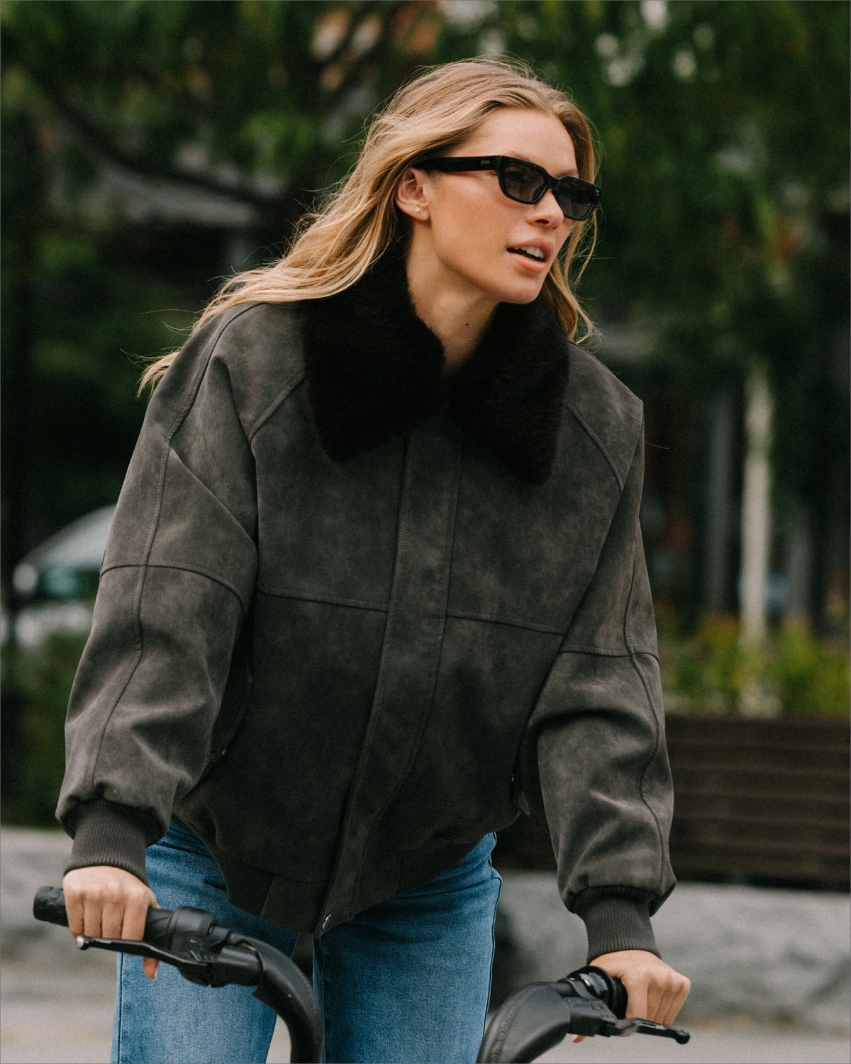 A woman riding a bicycle outdoors wearing a dark charcoal gray suede jacket with a black faux-fur collar and blue jeans.