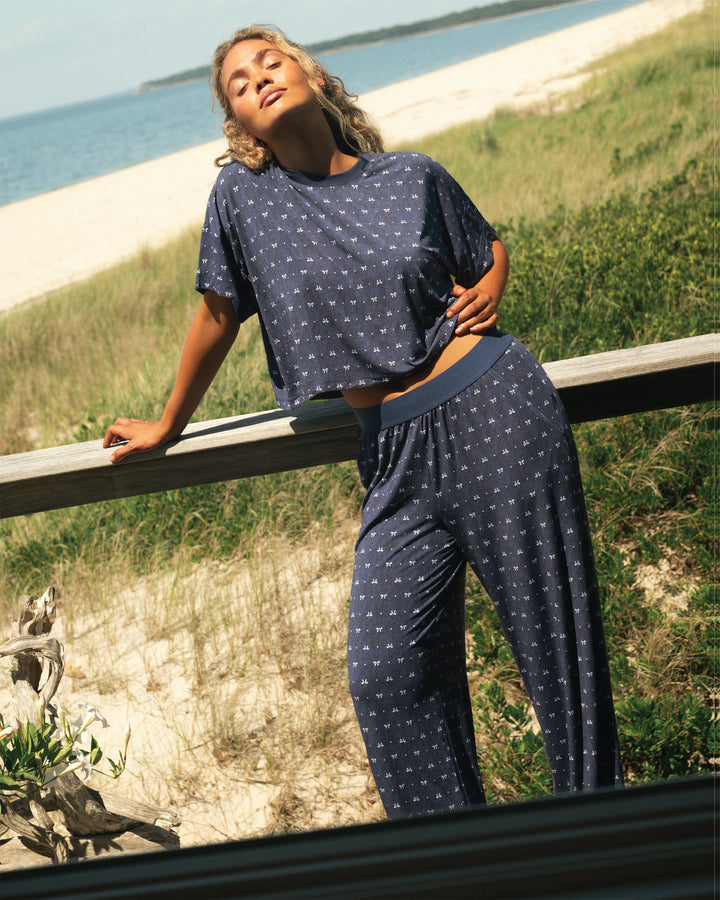 Woman in a blue polka dot matching pajama set standing by a wooden railing with a scenic beach background.
